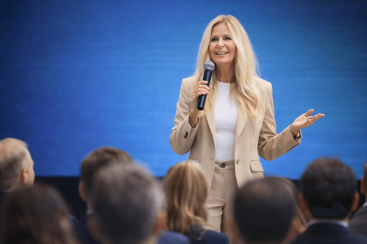 Lächelnde blonde Rednerin im beigen Hosenanzug hält eine Rede vor Publikum auf blauem Hintergrund.
