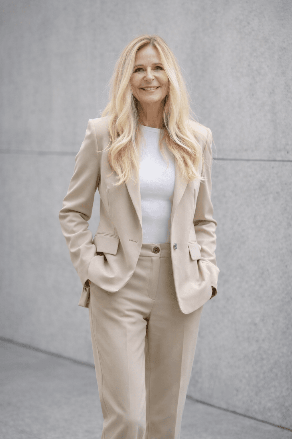 Lächelnde blonde Frau in elegantem beigem Anzug steht vor einer modernen grauen Betonwand.